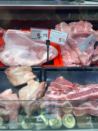 The image shows a close-up of a display case filled with various cuts of raw pork in a butcher shop. The lighting is bright, illuminating the fresh meat. In the foreground, on the left, there are several pieces of pork, some with skin and fat. To the right of these, a large cut of pork is visible, with a label indicating "E. JAMON CERDO C.P." and a price of "5,75 €/Kg". Further to the right, another cut of pork, identified as "Filete Jamon Cerdo V.a.", is priced at "6,30 €/Kg". In the background, more raw pork cuts are displayed. Below the meat, in the lower part of the image, metallic cans are visible through a glass partition. The overall environment appears to be a commercial setting for selling meat products. The location is specified as Bel Air, Spain.