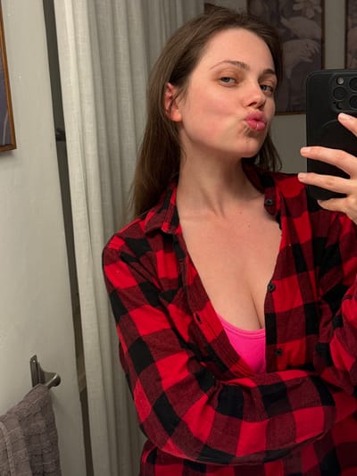 A woman with brown hair is taking a selfie in a bathroom mirror. She is wearing a red and black plaid shirt over a pink top. Her lips are pursed in a kissing gesture. The bathroom features light-colored walls, a towel rack with a gray towel, and blinds on a window. Part of a framed picture is visible in the bottom left corner. The scene appears to be indoors, and there is no indication of it being in Perugia, Italy.