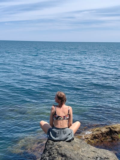 A woman with braided hair sits cross-legged on a rocky shore, facing the ocean in Utios, Ukraine. She wears a floral bikini top and grey shorts. The sea is calm, with gentle waves lapping at the shore. The sky above is clear blue with scattered white clouds. The sun is shining brightly, casting a warm glow on the scene. The woman appears to be enjoying the peaceful atmosphere and the beautiful natural scenery.