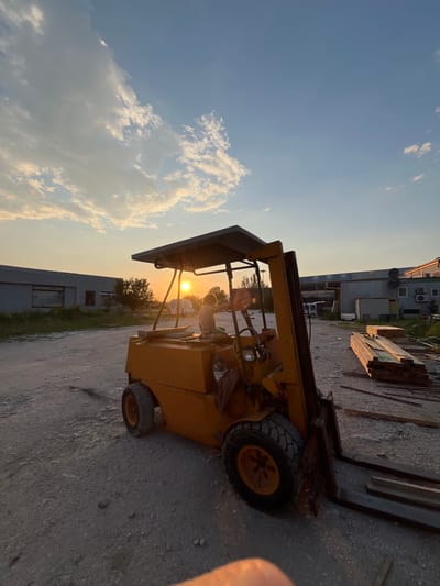 The image shows a yellow forklift parked on a paved surface in what appears to be an industrial or construction area in Pobrdje, Montenegro. The sun is setting, casting a warm glow across the scene. A child is seated in the forklift's driver seat. In the background, there are buildings, trees, and stacks of construction materials. The sky is visible with some clouds.