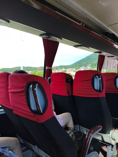 The image shows the interior of a bus with red seat covers, viewed from a seat behind the front seats. The seats are covered in red fabric with black lower sections. Each seat back has a circular opening with a clear plastic cover, through which a reflective surface is visible. The window behind the seats offers a view of a green, tree-covered hillside with scattered buildings. A red curtain is visible on the right side of the image. Above the seats, there are overhead compartments and lighting fixtures. The number "13 14" is visible on a panel above the windows. A sign with text in Cyrillic script, possibly "ИНФОРМАЦИЯ," is on the right side of the bus. A person's legs, wearing light-colored shorts with a strawberry pattern, are visible in the foreground on the right. A dark green purse rests on the seat next to them. The location is identified as Agoy, Russia.