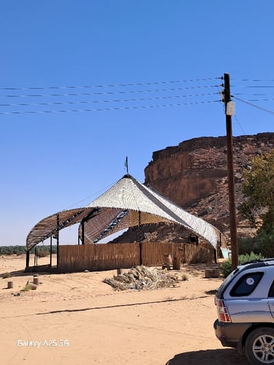 The image shows a scene in Karima, Sudan, at the coordinates 18.54 latitude and 31.831 longitude. A large, white, open-air structure with a curved roof is situated on a sandy lot. The structure is made of wood and has a woven fabric roof. A tall wooden pole with several electrical wires running across it is located to the right of the structure. There is a rocky cliff in the background behind the structure. There is also a silver SUV parked on the sandy lot to the right of the structure.  The SUV has a sticker on the back window. The sky is clear and blue. There are no people visible in the image.  The environment is arid and dusty. The scene is likely a market or a place where people gather.