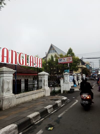 This image depicts an overcast street scene in Chagzam_Pam, Bhutan. In the left foreground, a large red and white sign displays the word "ANGLAYA" above a building with a brown tiled roof and a white, classical-style fence featuring black decorative railings and substantial white pillars. Adjacent to this fence, a grey sidewalk runs parallel to a wet asphalt street. The street features a black and white striped curb and white lane markings. A wrapper of litter lies on the wet street surface. A person wearing a dark jacket and helmet is riding a light-colored scooter away from the viewer, with its right turn signal illuminated. Further down the street, other motorcycles are visible. In the background, behind the "ANGLAYA" building and trees, a larger building with a steep, grey-pitched roof is prominent. A red and white billboard for "POS IND," "POSPAY," and "Pos Aja!" stands in front of this building. The sky is uniformly light grey and overcast.