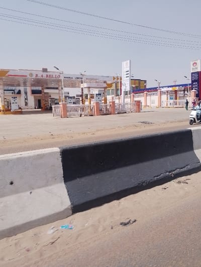 The image displays an exterior scene in Chiranchi, Nigeria, under a clear, pale blue sky. In the immediate foreground, a concrete road median barrier is visible, painted white on its upper section and black on its lower half. Directly in front of the barrier lies a sandy dirt shoulder with scattered debris, including a blue plastic wrapper.
The midground transitions into a paved area leading to a large commercial complex. On the right side of this paved area, two individuals are seen riding a white scooter-style motorcycle. Further into the complex, a solitary individual in dark clothing is observed walking near a perimeter fence.
The background is occupied by two adjacent fuel stations. The station on the left is identified by overhead signage as "B.A. BELLO NIGERIA LIMITED" and features multiple fuel pumps under a white canopy with yellow/gold accents. A building with a dark roof and light-colored walls stands behind the pumps, displaying a "DIESEL" sign. The station on the right, indicated by prominent vertical signage, bears the "SALBAS" brand and also presents fuel pumps beneath a canopy. This "SALBAS" sign lists "DIESEL," "PMS," "Kerosene," "LPG," and "Lubricant" as available products. White and pink/peach-colored fencing and bollards delineate the boundaries and entrances of both fuel stations. Multiple thin power lines are visible stretching across the upper portion of the frame. The ground throughout the scene appears generally dry and dusty.