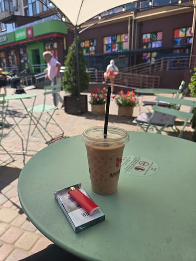 An outdoor cafe scene in Petropavlivska Borshchahivka, Ukraine features a light green, round table in the foreground. On the table rests a clear plastic cup with a beige beverage and a black straw, decorated with red triangles. Next to the cup is a pack of white and green "Winston Superline" cigarettes, topped with a red disposable lighter. In the background, people are visible walking near buildings with green and brown facades. One building displays colorful square artwork. Several light green metal chairs and tables are scattered throughout the area, and potted plants with red flowers add a touch of color. The ground is paved with interlocking bricks.