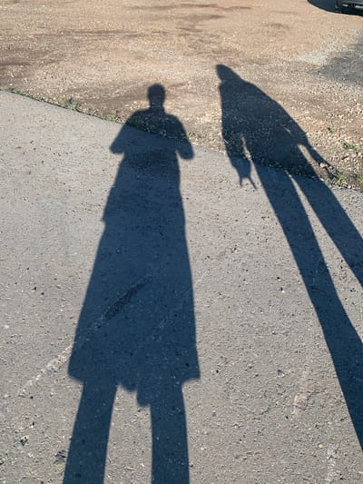 Two long, dark shadows are cast on an asphalt surface, indicating a sunny day. The shadow on the left depicts a person wearing a long coat or dress, with one arm extended to the side. The shadow on the right shows another person with their arm raised, possibly in a greeting or gesture. The ground transitions from asphalt to a gravelly area with sparse vegetation along the edge. A portion of a dark-colored vehicle is visible in the upper right corner of the image, suggesting it is parked nearby. The overall scene appears to be an outdoor public space in Verkhnii Sekines', Russia.