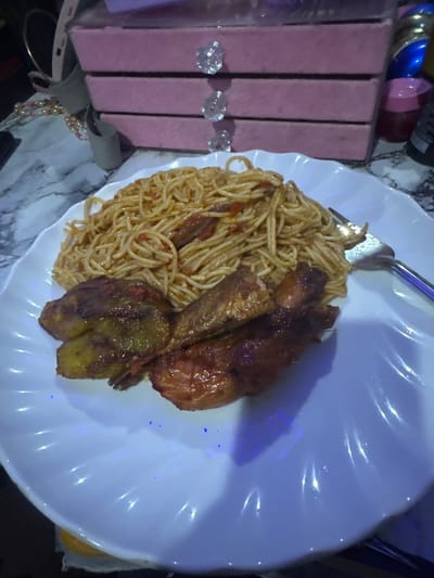 A close-up, overhead shot reveals a meal served on a white, wavy-edged plate, resting on a marble-patterned surface. The main subjects are a portion of spaghetti mixed with a light, possibly tomato-based sauce, and several pieces of what appear to be fried or roasted plantain and chicken, all with a dark, glossy, golden-brown finish. A fork or spoon is partially visible at the top of the plate, resting on the spaghetti.
In the background, to the left, are three stacked, rectangular boxes or containers in a muted pink or mauve color. Each box features a clear, diamond-shaped handle or knob. Behind these boxes, there are glimpses of other metallic or reflective objects, including blue, gold, and maroon items. A string of light-colored beads is also visible near the bottom left, resting on the marble surface.
The scene is indoors, likely a dining or kitchen setting. The lighting is somewhat dim, with prominent blue and purple reflections visible on the white plate, possibly from artificial light sources. No people are visible, and no text is discernible in the image. The food presentation suggests a prepared meal.