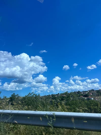 The image depicts a scenic view of a hillside village under a bright blue sky dotted with fluffy white clouds. The foreground features a grey metallic guardrail running horizontally across the frame, with dry grass and vegetation blurred in front of it. Behind the guardrail, a lush green hillside is covered with trees and bushes. Scattered across the hillside are numerous houses with red-tiled roofs, characteristic of the architecture found in Konstantinovo, Bulgaria. The scene suggests a rural or semi-rural setting on a clear day.