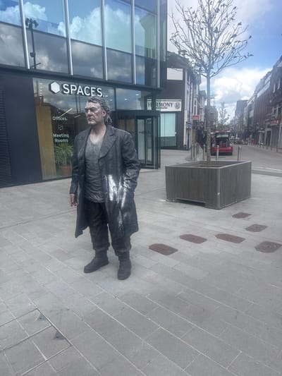This image captures a pedestrianized urban scene in Woking, United Kingdom, specifically near Block E, The Lightbox, Dukes Walk.
In the foreground, a male street performer, or "living statue," stands facing towards the viewer's right. He is covered head-to-toe in metallic grey-bronze body paint, including his face, hands, and clothing. He wears a long, dark overcoat, a dark t-shirt, and dark trousers, all appearing to be painted to mimic aged metal or stone. Visible white paint streaks are present on his left arm and the front of his overcoat. His expression is neutral.
To the left of the performer is a modern building with a dark facade and large, reflective glass windows that reflect a partly cloudy sky. The ground floor displays a prominent white "SPACES." logo, with smaller "Offices" and "Meeting Rooms" text below. A revolving glass door serves as an entrance.
To the right of the performer, and extending into the background, is a wide, paved street. A bare tree, featuring dark, sparse branches and small, dark ornamental leaves, is planted within a large, dark grey rectangular planter. Further down the street, older brick and stone buildings line both sides, one displaying a "HARMONY" sign. A red vehicle with an open tailgate is parked in the distance. The ground is paved with light grey rectangular flagstones, interspersed with various rectangular and square dark metal utility covers. The sky is partly cloudy.
