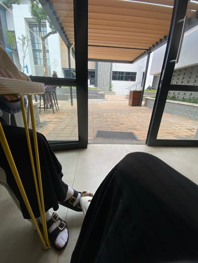 The image displays a scene in Sandton, South Africa, captured from an interior vantage point looking out into an outdoor courtyard. In the immediate foreground, the legs of the photographer, clad in dark trousers, are visible on the right. To the left, another individual's legs and feet are visible, wearing black trousers, white socks, and dark brown two-strap sandals with gold-colored buckles. A light-colored table with thin yellow vertical supports stands between the two individuals.
The interior space features light-colored tiled flooring and is separated from the exterior by a dark-framed glass doorway, which appears to be open. A dark rectangular doormat is situated directly outside the doorway on the paved ground.
The outdoor courtyard is paved with reddish-brown interlocking pavers. A dark-framed pergola with a light brown slatted roof extends over a portion of the courtyard. Modern buildings with white and grey facades, featuring large windows, define the perimeter. A large blue numeral "1" is visible on the white facade of a building on the left, partially obscured by a slender tree. Multiple planter boxes, constructed from grey block-like material, contain green foliage, and a dark wooden planter box is centrally located.
Two individuals are visible in the courtyard. One person is seated at a high table with two dark stools in the left mid-ground. A second person is faintly visible in the far right mid-ground, near additional raised garden beds and a white building facade. The scene is illuminated by natural daylight.