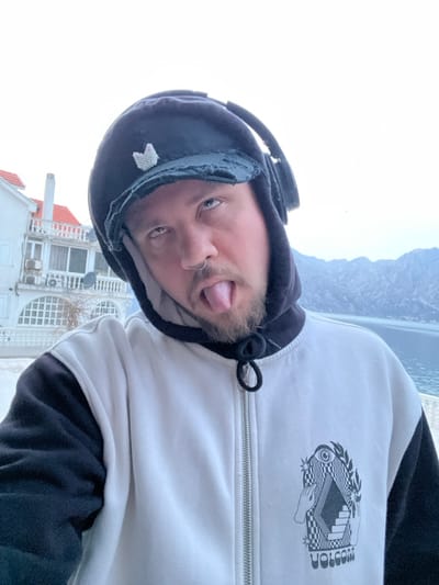 A male individual is depicted in a chest-up, outdoor selfie, against the backdrop of Bogdašići, Montenegro. He has a beard and mustache, a ring in his left nostril, and is sticking his tongue out while looking upwards. He wears a black hoodie with the hood pulled up, over which a black, distressed baseball cap with a small, light-colored butterfly-shaped pin is worn. Black over-ear headphones rest on his ears over the cap. He is dressed in a two-toned zip-up jacket, primarily white on the torso with black sleeves, featuring a prominent graphic design on the right chest area that includes an eye, hands, geometric patterns, and the "VOLCOM" logo.
The background shows a white, multi-story building with a red-tiled roof, multiple balconies, and external air conditioning units on the left. To the right, a body of water (likely the Bay of Kotor) is visible, flanked by steep, rocky mountains in the distance under an overcast sky. The individual appears to be positioned near a light-colored paved area, possibly a promenade or balcony overlooking the bay.
