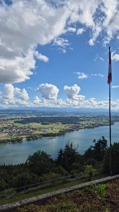 This image presents a panoramic view from an elevated position overlooking Lake Biel/Bienne and its surrounding landscape in the region of Biel/Bienne, Switzerland. The scene is observed under a bright blue sky with scattered white cumulus clouds of various sizes.

In the immediate foreground, a low, weathered stone wall spans horizontally across the lower portion of the frame, partially obscured by dark green ground cover and low vegetation. Behind this wall, a strip of grass is visible, followed by a chain-link fence, which is partially visible through further dense green foliage. These elements suggest a perimeter or viewing area.

The midground is dominated by the expansive, blue-green waters of Lake Biel/Bienne, extending from the left to the right side of the frame. The lake's surface exhibits minor ripples and reflections under bright sunlight. Several small, white-hulled boats are visible on the water, appearing as static objects. Along the far shore of the lake, a densely forested area transitions into an urbanized landscape, identifiable as a segment of Biel/Bienne or a neighboring municipality. This area features numerous buildings with varied roof colors, primarily red and grey, interspersed with green parks and fields. Further beyond, agricultural lands and additional forests are visible.

The background includes distant, hazy blue mountain ranges, likely the Jura mountains, on the horizon. On the far right, a tall, dark flagpole stands vertically, extending from the midground into the sky. Attached near its top is a flag, unfurled by an unseen breeze, displaying a red field with a distinct white section, characteristic of the national flag of Switzerland. No individuals, explicit actions, or direct interactions are discernible within the frame.
