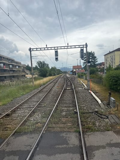 The image depicts a railway junction in Reconvilier, Switzerland, under a cloudy sky. Multiple sets of train tracks converge and diverge, with gravel and grass lining the ballast. Overhead power lines stretch across the scene, supported by metal gantries. A railway signal, displaying a vertical row of three lights, is visible on the left side of the image, with another similar signal on the right. In the background, apartment buildings and houses line the left and right sides of the tracks, respectively, interspersed with trees and vegetation. The scene appears to be a functional train station or a point of railway infrastructure. No people are visible in the image.