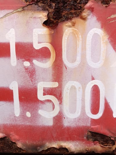 The image is a close-up, weathered view of white numbers spray-painted on a red surface, likely metal. The numbers appear to be "1.500" repeated twice, with a rust-colored edge along the top and bottom. The paint shows signs of cracking and wear. There is no indication of the city of Blangbladeh, Indonesia, or any people in the image.