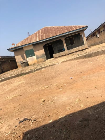 The image displays a single-story building in Okinni, Nigeria, situated across an unpaved dirt road. The building features a weathered, corrugated metal roof with a light blue trim and light-colored walls showing discoloration and staining. A covered porch supported by two rounded pillars spans the front. An open doorway on the porch reveals a dark interior, and an adjacent window frame is open without glass. A blue and white striped object, possibly a recliner, is visible inside the porch. To the left, a window with green shutters or frames is present, alongside a well with a concrete rim. A blue and white satellite dish is mounted on the left side of the roof's overhang. The foreground consists of a reddish-brown dirt road with scattered dry vegetation and minor debris, with a shadow cast across the lower right. A low concrete block wall borders the property to the left, and a partial view of another concrete block building is visible to the far right in the background. The sky is clear blue. No individuals are depicted.