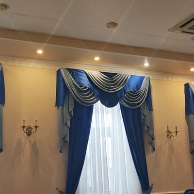 This image captures an interior scene, likely a formal room such as a dining hall or event space in Novo-Nikol'skoe, Russia. The primary subjects are the elaborate window treatments and the room's elegant decor.

A large window, covered by sheer white curtains, is flanked by ornate, two-tone drapes. The drapes feature a royal blue fabric combined with a lighter grey-blue, arranged in decorative swags and valances at the top, and flowing panels on the sides. Two antique-style, brass-colored wall sconces with crystal embellishments and electric candle bulbs are mounted on the light beige walls, one on each side of the central window. These sconces cast prominent, elongated shadows on the wall, suggesting illumination from the front.

The ceiling above is light-colored, possibly a suspended ceiling with acoustic tiles, and features several recessed spotlights that provide additional artificial illumination. A circular device, possibly a smoke detector, and an air conditioning vent are also visible on the ceiling. Decorative crown molding with a swirling pattern runs along the top of the walls, just below the ceiling.

The scene appears static with no people or ongoing activity. The artificial lighting and formal furnishings suggest an indoor setting designed for gatherings or events. No text is visible in the image.