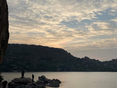 The scene depicts a natural landscape at Sanapur Lake in Sanapur, India, during either dawn or dusk. A calm body of water, likely a lake, occupies the foreground and middle ground, reflecting soft light. Several large, rounded grey boulders protrude from the water in the immediate foreground. A dark, rocky hill range, sparsely covered with vegetation, spans the mid-ground, silhouetted against the sky. A large, dark rock face partially frames the left side of the image. The sky is largely covered by altocumulus clouds, displaying light blue-grey tones with soft yellow and faint orange hues near the horizon. A small, distinct patch of pinkish-red light is visible above the distant hills on the left, indicating the sun's position. Three individuals are present: two are clearly visible as dark silhouettes standing on separate boulders in the foreground. One stands upright, facing generally right, while the other is slightly hunched over, also facing right. The head and shoulders of a third individual are partially visible and heavily cropped in the bottom-left corner, silhouetted against the water. The individuals are positioned on the rocks overlooking the water.