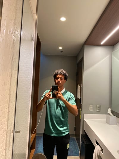 A person with curly hair holds a phone in a bathroom mirror, capturing a selfie. The individual wears a green t-shirt, a white collar, and black pants with a logo. The bathroom features white walls, recessed lighting, and a horizontal light fixture. The mirror reflects a tiled shower, while the background shows a glimpse of a blue carpet. A tissue box and light switches are visible above the sink.