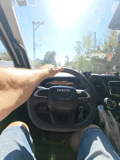 The image shows a first-person perspective of someone driving a vehicle. The driver's left arm, covered in fine hairs, is visible on the steering wheel, gripping it to navigate. They are wearing denim shorts. The steering wheel has the "IVECO" logo prominently displayed in the center.

The scene is set inside the car, looking forward through the windshield. The dashboard is visible, with a gear shifter on the right side showing positions R, N, D, and a manual mode indicator. A small skull figurine is attached to the dashboard near the top right, and other items are present on the dashboard, including what appears to be a phone mount and possibly some cables.

Outside the car, the weather appears to be sunny, with bright sunlight creating lens flare and washing out some of the details. The sky is clear blue. Trees and buildings are visible in the distance, suggesting an outdoor, daytime setting. A utility pole with wires is also visible against the sky.

On the windshield, near the top left, there are stickers. One sticker has Hebrew text and a phone number, "03-9225310". Another sticker also contains Hebrew text.

The overall impression is of a person driving during a bright, sunny day, possibly in a suburban or rural area given the vegetation and relatively clear road ahead. The image captures a moment of everyday activity, the act of driving. The sunlight suggests it is likely late morning or afternoon.