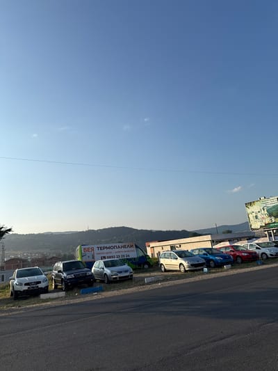 The image shows a row of cars parked on a grassy incline next to a road. In the background, there are hills covered with trees and a few buildings. A truck with an advertisement is parked behind the cars. A billboard for organic products is visible on the right side of the image. The sky is clear and blue with a few faint clouds. The sun is setting, casting long shadows. The location is identified as Kraishte, Bulgaria.