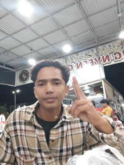 A young man, likely in his early twenties, is captured in a selfie taken outdoors at night. He is wearing a plaid collared shirt over a dark t-shirt. His right hand is raised, with his index and middle fingers extended in a V-shape, positioned near his face. The background reveals a covered outdoor seating area with a corrugated metal roof illuminated by fluorescent lights. A large sign with red and yellow lettering, seemingly in Indonesian, is visible behind him, possibly indicating a food establishment. Further in the background, other individuals are present, including a person in a yellow shirt. The scene appears to be in a casual, public setting, possibly a street food area or outdoor market, likely within the city of Sigli, Indonesia, given the context provided.