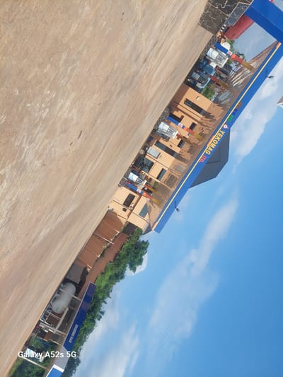 The image shows a gas station in Ogbunike, Nigeria, with a blue and yellow canopy over the pumps. The building is an orange-brown color, and there are trees and a clear blue sky in the background. A white gas tank is visible on the left side of the image. The text "YEKOMAG LTD." is visible on the canopy, and "COOKING GAS" is visible on a blue sign. A "Galaxy A52s 5G" watermark is present in the bottom left corner of the image.