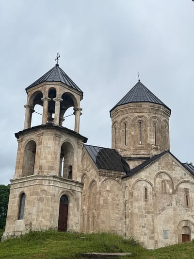 The media file shows a close-up, vertically oriented view of a stone church structure, likely the Nikortsminda Cathedral in Georgia. The main subjects are architectural elements of the church, specifically a dome and a bell tower, both constructed from light-colored stone with dark, pitched roofs. Intricate carvings and arched windows are visible on the tower and the main body of the church. One of the domes is topped with a cross, and the bell tower contains visible bells.
The scene is set outdoors, with a gray, overcast sky suggesting either early morning or a cloudy day. To the right of the church, a patch of lush green grass slopes upwards, indicating a natural setting. The stone appears weathered, suggesting the age of the structure. There is a dark wooden door visible on the side of the church. No people are present in the frame, and no text is discernible. The overall impression is one of historical grandeur and a somber atmosphere due to the overcast weather.