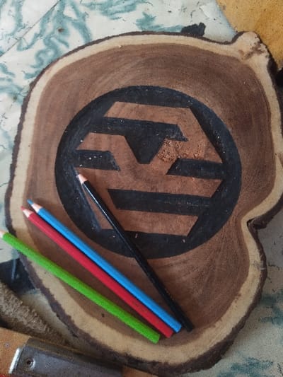 A irregularly shaped wood slice, approximately 20-25cm in diameter, rests on a light-colored surface with a swirling green and white pattern, possibly a cloth or a table covering. The wood slice features a geometric, stylized hexagonal design etched into its center, partially filled with matte black pigment. Visible within the design are traces of reddish-brown material, possibly wood dust or pigment. Four colored pencils are positioned diagonally across the lower left portion of the wood slice: one black, one blue, one red, and one green. A metallic tool with a light brown wooden handle is partially visible at the bottom left corner of the frame. The scene is static, implying a workbench or crafting area in Жарсуат, Kazakhstan. No individuals are present.
