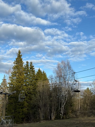 The image shows a ski lift ascending a wooded slope under a blue sky with scattered clouds. The ski lift is the dominant feature, with empty chairs suspended from cables stretching across the frame. Below the lift, a mix of evergreen and deciduous trees covers the hillside. The ground is visible along the right edge of the frame, appearing as a grassy area. The location cannot be definitively determined to be Galevo, Russia based on the image alone.