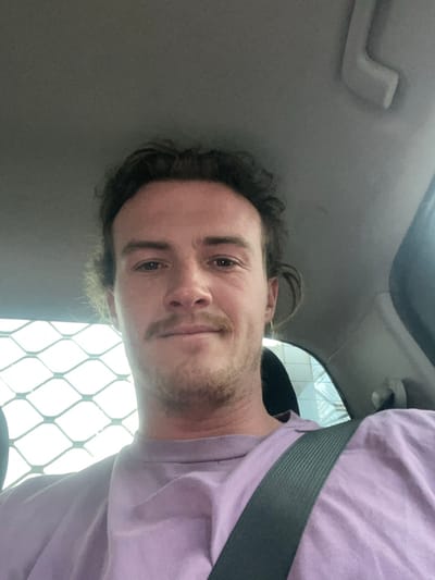 A male individual is depicted in a front-facing mid-shot within the interior of a vehicle. He appears to be in his 20s to 30s, with light skin, dark wavy hair, and a mustache. He is wearing a light purple crew-neck t-shirt and has a dark seatbelt fastened across his left shoulder and chest. The individual looks directly at the camera with a slight smile. The vehicle's light-colored headliner and a grab handle are visible above him. Behind the subject, through a rear window on the left, a light-colored metal grid or security mesh is visible, suggesting a utility or cargo area. On the right, through another window area, indistinct light-toned architectural elements are visible in the background. The specific location within Thirroul, Australia, cannot be determined from the visual information provided.