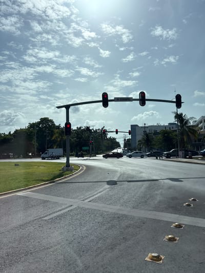 The image captures a street scene in Cancún, Mexico, specifically at the intersection of Avenida Bonampak. The sky is partly cloudy with bright sunlight, casting shadows on the asphalt road. Traffic lights are prominently displayed, with the lights appearing red, indicating a stop. Several vehicles are present, including cars, a white van, and a motorcycle. One person is riding the motorcycle. The road shows clear lane markings and a crosswalk. The surrounding environment features green grass, trees, palm trees, and buildings. The overall impression is of a typical urban intersection on a sunny day in Cancún.