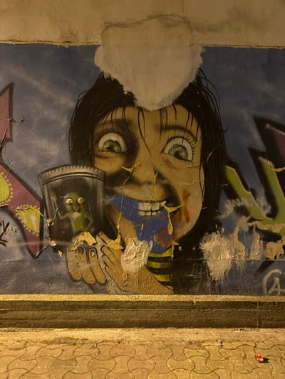 An outdoor concrete wall in Giannitsa, Greece, features a large, defaced graffiti mural under artificial yellow light at night. The central subject of the mural is a cartoonish figure with dark hair, wide green eyes, and a broad, toothy grin. This figure, depicted with light brown skin and wearing a yellow and black striped top, holds a transparent jar containing a small, green, alien-like creature with large black eyes and three-fingered hands, one of which is raised. The mural's background is primarily blue, depicting a sky with partial white cloud shapes, and includes elements of other graffiti in pink, purple, and green. Significant paint and plaster damage is evident across the mural, with large areas of white flaking and peeling exposing darker layers beneath, notably over the main figure's head and mouth. Below the wall, a raised concrete curb separates it from a paved sidewalk with a light brown, interlocking, rounded pattern. A small red piece of debris is visible on the sidewalk in the lower right quadrant.