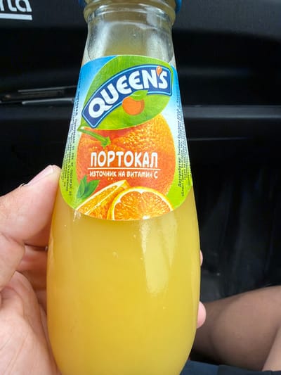A close-up shot shows a glass bottle of "QUEEN'S" brand orange juice held by a person's hand. The label features the brand name in large, white, outlined letters against a blue and green background. An illustration of an orange and orange slices is also visible. Text on the label, in Bulgarian, reads "ПОРТОКАЛ" (Portokal - Orange) and "ИЗТОЧНИК НА ВИТАМИН C" (Source of Vitamin C). To the right of the main label, there is a smaller text block with distribution information, including "Дистрибутор: Тимбарк България ЕООД, с. Казaнчеев..." (Distributor: Timbark Bulgaria EOOD, s. Kazancheev...). This suggests a location in Bulgaria, possibly near the village of Kazancheevo. The background is dark and out of focus, indicating an indoor or shaded environment. The overall impression is a product shot focused on the orange juice bottle.