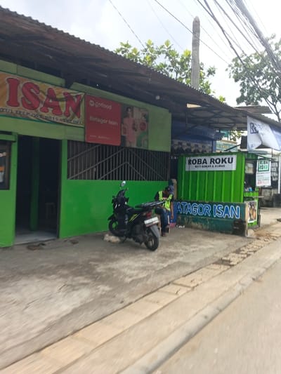 The scene depicts a street-level view of commercial establishments in Margahayu, Indonesia. On the left, a single-story building with a green facade and a corrugated metal roof features a prominent red banner displaying "ISAN," the "gofood" logo, and "Batagor Isan," accompanied by an image of a woman. To the right of this building, a smaller green kiosk or stall is visible, bearing the text "ROBA ROKU" and "ROTI BAKAR & KUKUS." Further right, another stall with a blue awning is partially visible. A black scooter/motorcycle with a rear luggage rack is parked on the concrete sidewalk in front of the green building. A male individual, wearing a dark cap, a bright yellow safety vest, and dark trousers, is seated on a low structure next to the "ROBA ROKU" stall, facing generally away from the camera. Overhead, numerous utility lines traverse the cloudy sky, and tree foliage is visible above and behind the structures. The foreground consists of a cracked concrete sidewalk transitioning to an asphalt road.