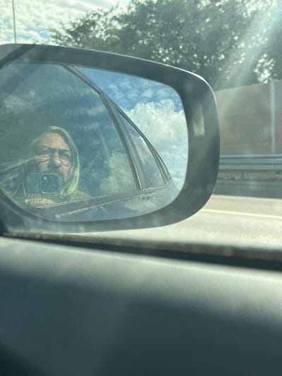 A daytime image captured from inside a vehicle shows the reflection of an individual in the passenger-side door mirror. The individual, appearing to be male, has long light-colored hair, a full beard, and wears glasses with dark frames. They are holding a dark-colored smartphone, equipped with visible camera lenses, up to the mirror, indicating they are taking the photograph. Faintly visible text, "OBJECTS IN MIRROR ARE CLOSER THAN THEY APPEAR," is present on the mirror's surface. The mirror also reflects a blue sky with scattered white clouds, and parts of the vehicle's interior window frame. Outside the mirror, a multi-lane asphalt road or highway is visible, running alongside a tall, light-colored (tan/gray) sound barrier or wall. Beyond the barrier, blurred green trees are present. Sunlight is bright, evidenced by a lens flare in the upper right area outside the mirror. The immediate foreground shows a dark gray interior door panel of the vehicle. There are no identifiable landmarks within the image to specify a location within the Sdot Dan Regional Council, Israel.