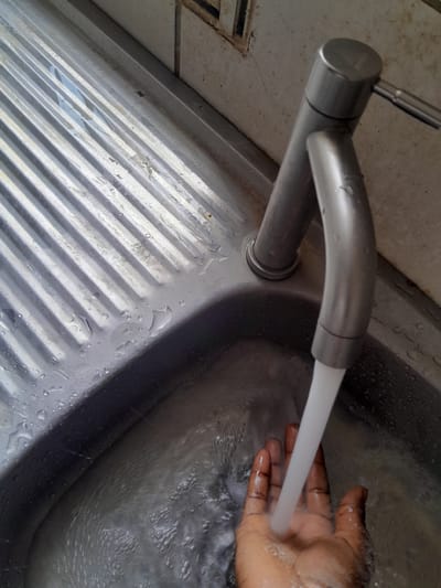 The image shows a close-up, overhead view of a person's hand being washed under a faucet in a sink. The hand is dark-skinned and appears to be an adult's. Water is flowing from a modern, metallic faucet into the sink, which is also made of metal and has a ribbed texture. The background consists of a wall or countertop area that appears to be made of a light-colored, textured material, possibly concrete or stone. There are some water droplets visible on the surfaces. The lighting suggests it is daytime. The scene is likely taking place indoors, in a bathroom or kitchen. There is no visible text.