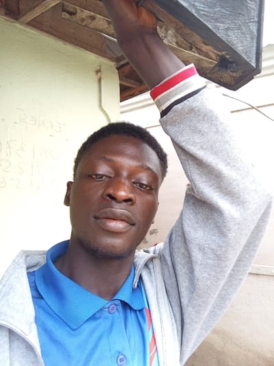 A young Black male is pictured in a close-up, selfie-style photograph taken outdoors. He is wearing a blue collared shirt with two purple buttons and a gray hooded jacket. The jacket has a red, white, and black striped band around the cuffs. The subject's left arm is raised, holding onto a dark wooden beam that appears to be part of an overhead structure. The background consists of a textured, light-colored wall with faint writing on it, and the wooden structure above. The setting suggests an outdoor, possibly residential or communal area in Gindin Dutse, Nigeria. The lighting is natural and appears to be daylight.