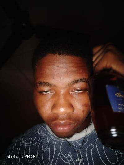 This is a close-up, waist-up, selfie-style photograph of a young Black male, possibly in his late teens or early twenties, holding a bottle of alcohol to his right cheek and ear. The individual has short, dark, coiled hair and his eyes are partially closed, with his head tilted slightly back. His skin appears to be moist, possibly due to sweat, and there are visible blemishes on his forehead and cheeks. He is wearing a dark, long-sleeved shirt with a white swirling pattern. A silver cross pendant is visible on a chain around his neck. The background is dark and out of focus, with what appears to be a ceiling fan visible in the upper left corner. The text "Shot on OPPO R11" is present in the lower left corner, indicating the device used for capture. The label on the bottle is partially visible, with the words "Aged", "DVM", and "ALCO" discernible. The image appears to have been taken indoors, likely in a residential setting, and the lighting is dim. The subject's location in Akokwa, Nigeria, is inferred from the context provided.