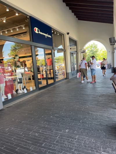 The image shows the exterior of a Champion store in an outdoor mall setting, possibly an outlet mall, as suggested by the presence of multiple brand stores and the architectural style. The Champion store has a large glass storefront displaying mannequins dressed in athletic wear and sale signs. Next to the Champion store is another branded store, possibly Puma, indicated by a hanging sign. Several people are visible walking along the covered walkway in front of the stores, some carrying shopping bags. The walkway is paved with dark, rectangular stones. The architecture includes arched openings and a wooden-beamed ceiling. The scene appears to be in Rodengo, Italy, as indicated by the context provided. The lighting suggests it is daytime, with sunlight filtering through the trees and the open areas of the mall.