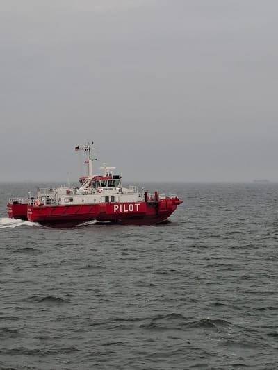 A red and white pilot boat with the word "PILOT" in large white letters on its side is traveling through the water. The water is gray and choppy. The sky is overcast and gray. The pilot boat is in the city of Cuxhaven, Germany.