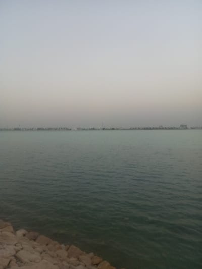 The image shows a view of the waterfront in Al Khobar, Saudi Arabia. A body of water, likely the Persian Gulf, occupies the majority of the frame. The water appears calm with subtle ripples. In the distance, a faint skyline of buildings and structures can be seen along the coastline. The sky is overcast with a soft, diffused light. In the foreground, there is a rocky shoreline. There are no people visible in the image.