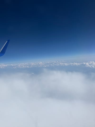 The image shows an aerial view from an airplane window, looking out at a vast expanse of clouds and a deep blue sky. An airplane wing, painted blue and white, is visible in the upper left corner of the frame. The clouds are dense and appear to be a thick layer, obscuring whatever is below. The sky transitions from a lighter blue near the horizon to a darker, richer blue at the top of the frame. There are no visible structures or landmarks that would suggest a specific location within Karlovac, Croatia. The scene is one of serene isolation, high above the earth.