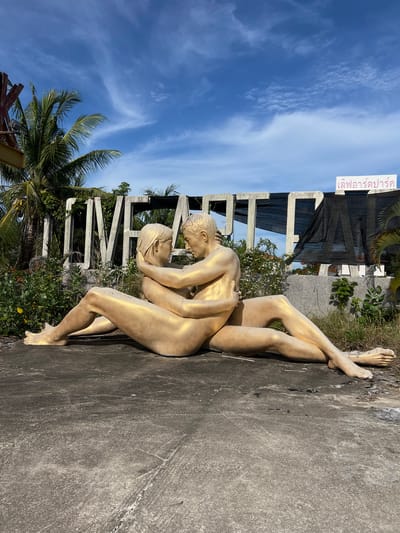 This media file shows a statue of two nude figures embracing, painted gold. The figures are positioned in a reclining pose against a concrete surface. Behind them, a large, partially obscured sign with large letters is visible, with greenery growing around and over it. A white sign with Thai text is also affixed to the top of this structure. To the left, palm trees and lush vegetation are visible under a bright blue sky with wispy white clouds. The sun appears to be shining, casting shadows and illuminating the scene, suggesting it is daytime. The overall setting seems to be outdoors, possibly in a tropical or subtropical location, given the vegetation and palm trees. The Thai text on the white sign translates to "Ban Noensai," which is likely the name of the location, "Ban Noensai, Thailand." The statue's gold color and the embrace of the figures might evoke themes of affection, art, or a memorial. The concrete surface shows signs of wear and tear.