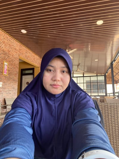A medium shot selfie features a young woman wearing a dark blue hijab and a dark blue long-sleeved shirt. She is seated outdoors at a cafe in Keraton, Indonesia. The background shows a brick wall on the left, a door with a sign that reads "201", and a ceiling with horizontal wooden panels. There are wicker chairs visible on the left and in the background. A ceiling fan is also present in the background. The woman is looking directly at the camera. The lighting appears to be natural daylight.