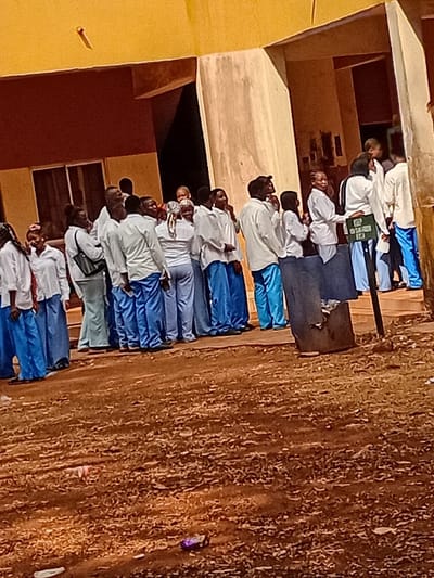 A group of people in school uniforms, consisting of white tops and blue bottoms, stand in a line, possibly for an event or assembly. They are positioned near a building with yellow and brown walls, with windows visible. The foreground shows a rough, dry ground covered in dirt and small debris. A sign is held by one of the individuals in the middle of the line. The scene has an outdoor setting with natural lighting.