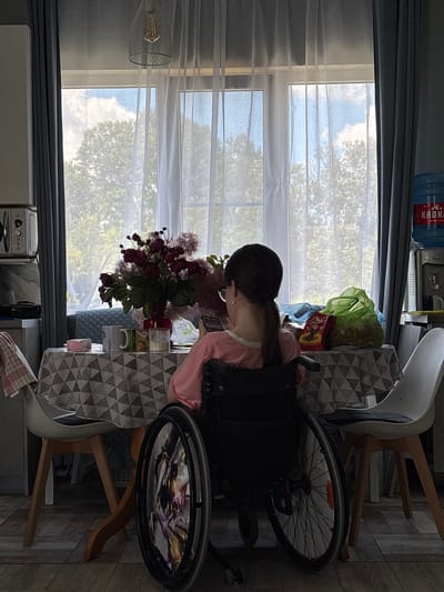A young woman with dark hair in a ponytail, wearing glasses and a pink t-shirt, sits in a wheelchair facing away from the camera. She is positioned at a table covered with a patterned tablecloth, where a red vase holds a bouquet of dark red flowers and fluffy pink-purple foliage. Two mugs and a small pink box are also on the table. Behind the woman, a large window is covered by sheer white curtains, revealing trees and a cloudy sky. To the right of the window, a blue water cooler with a red label and a dispenser is visible. The environment appears to be a dining area or kitchen within a home. The floor is made of wooden planks.