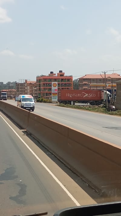 The image captures a daytime scene on a multi-lane highway in Sigona ward, Kenya, viewed from a vehicle. In the immediate foreground, a concrete median barrier with a tan hue runs diagonally across the lower left. The bottom edge reveals a dark gray dashboard and the vehicle's windshield frame.
On the paved highway, vehicles are in motion or stopped. In the left lane, a white semi-truck is followed by a white, blue, and orange passenger van displaying "NUCLEUR" on its side, both traveling away from the camera. On the right side of the road, a white semi-truck with a large red shipping container, marked "HAMBURG SÜD," is stopped or parked on the shoulder near a Shell gas station sign. A smaller, dark-colored sedan is partially visible behind this truck.
The background is dominated by several multi-story buildings. A prominent peach-colored building with multiple balconies and black rooftop water tanks is situated behind the Shell station. Further down the highway, other multi-story buildings with brown and grey facades are visible. The environment includes sparse roadside vegetation and utility poles with overhead lines on the right. The sky is a pale blue with a light haze and a few scattered white clouds. No specific individuals or direct human interactions are discernible within the frame.