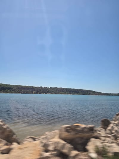 The image depicts a horizontal view of the Adriatic Sea and coastline near Tar, Croatia. The scene consists of a blue sky, a narrow strip of land densely covered in trees, and a large body of water with visible ripples. A rocky shoreline is blurred and prominent in the lower right foreground. The photograph is slightly out of focus, with a noticeable motion blur, especially in the rocky foreground. There are no people visible.