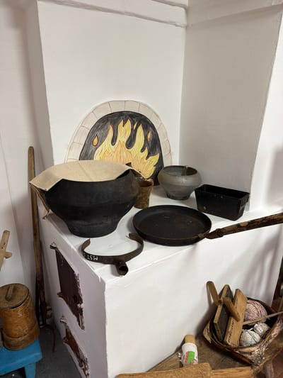 The image depicts a corner of a room with a white stove, likely a traditional Russian stove, against a white wall. The stove has a painted archway resembling a fire with flames. On top of the stove, there are several objects related to cooking and traditional crafts. A large, black, cast-iron pot, partially covered with a light-colored cloth, sits prominently in the foreground. To its right, there is a smaller, grey, metallic pot, and to the right of that, a black rectangular metal container. In front of the pots, a black frying pan with a wooden handle is placed on the stove's surface. A U-shaped metal clamp with white markings is also visible on the stove.
To the left of the stove, a tall wooden stick leans against the wall. Below the stove, there are two rusty metal doors, suggesting an oven or ash compartment. To the far left, a small blue wooden stool holds a cylindrical wooden container with a lid.
In the right foreground, a woven basket rests on a wooden surface. Inside the basket are several balls of yarn, various wooden tools, and what appears to be a bundle of natural fibers. A small doll-like object with a colorful fabric wrap is also present near the basket.
The scene suggests a historical or ethnographic display, possibly in a museum or a preserved traditional home. The location is identified as Ust'-Pyzas, Russia. There are no people visible in the image. The lighting is even, illuminating the objects clearly. The overall impression is one of rustic simplicity and historical preservation.