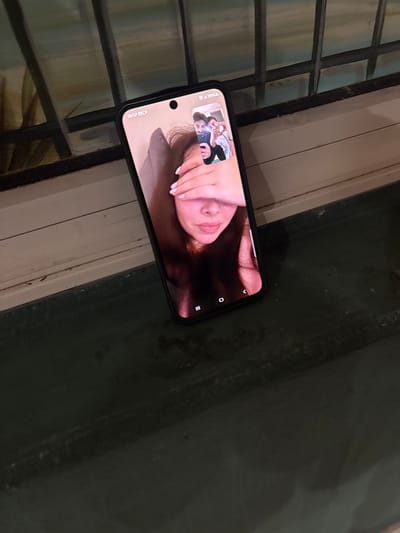 The image shows a smartphone positioned horizontally, displaying a video call in progress. On the main screen, a person with dark hair covers their eyes with a hand, revealing their nose, mouth, and chin. The hand has neatly painted pink fingernails. In a smaller picture-in-picture window at the top left, two other individuals, a young male and female, are visible, both smiling, suggesting a lighthearted interaction.

The smartphone is an Android device, featuring a punch-hole camera visible on the top left. The screen's status bar displays "THU 7.6" (possibly referring to Thursday, June 7th or 7:06) along with battery and signal icons. Partial text indicating "5G" and "2:30" is also visible below the punch-hole, likely denoting network status and call duration. Android navigation buttons are present on the bottom right of the screen.

The phone rests on a surface next to a window, through which a grid-like structure of bars or frames is visible. The external environment beyond the window appears dark and indistinct, suggesting an evening or nighttime setting. The scene takes place indoors, with the contextual location being Grenoble, France.